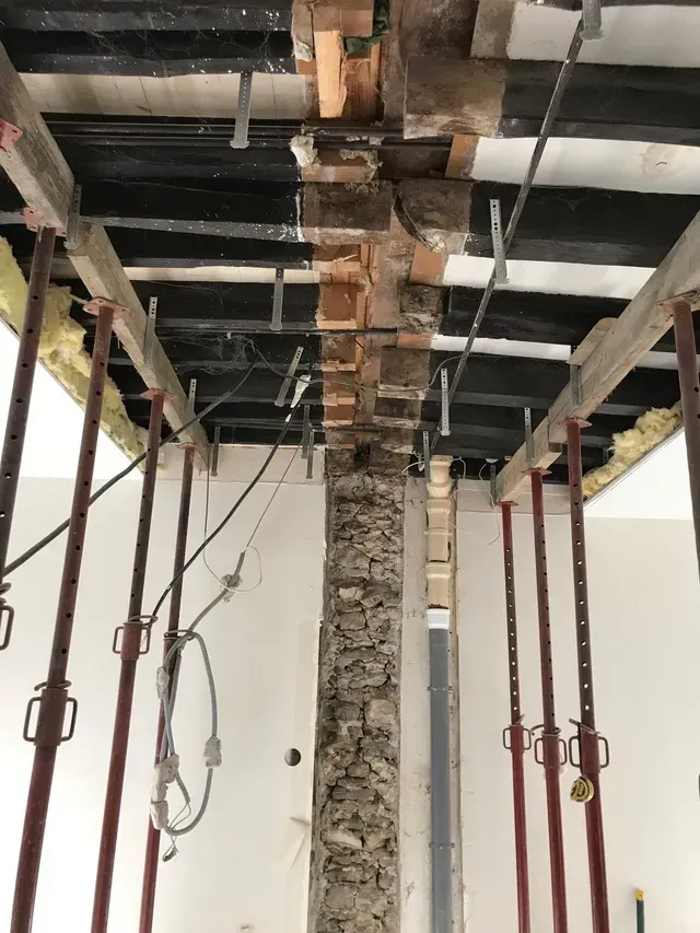 Chantier de construction avec poutres apparentes au plafond, une colonne centrale en pierre et des poteaux de soutien rouges.