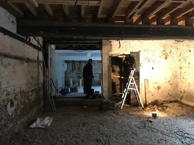 Pièce sombre et inachevée avec poutres apparentes, échelle et silhouette humaine se détachant sur le seuil.