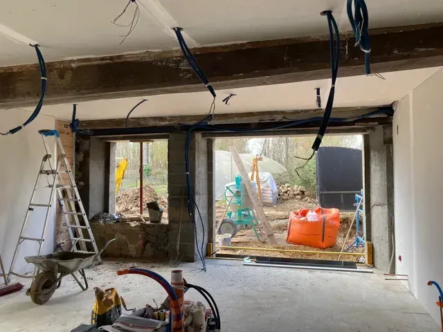 Chantier de rénovation avec poutres apparentes, échelles, outils et grande ouverture murale donnant sur une cour