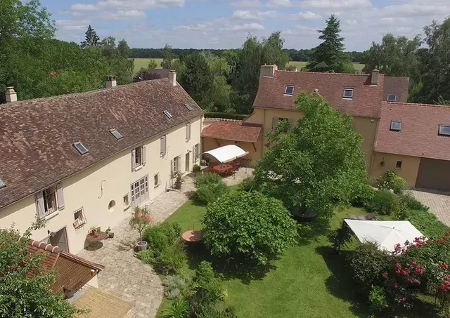 Cour rurale avec des maisons couleur crème, des toits de tuiles rouges, des arbres et des parasols blancs sous un ciel bleu