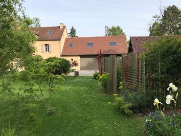 Jardin verdoyant avec pelouse et fleurs, menant à des maisons beiges aux toits rouges sous un ciel nuageux