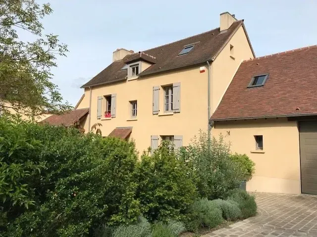Maison couleur crème avec un toit rouge, des lucarnes et un garage dissimulé derrière des haies vertes.
