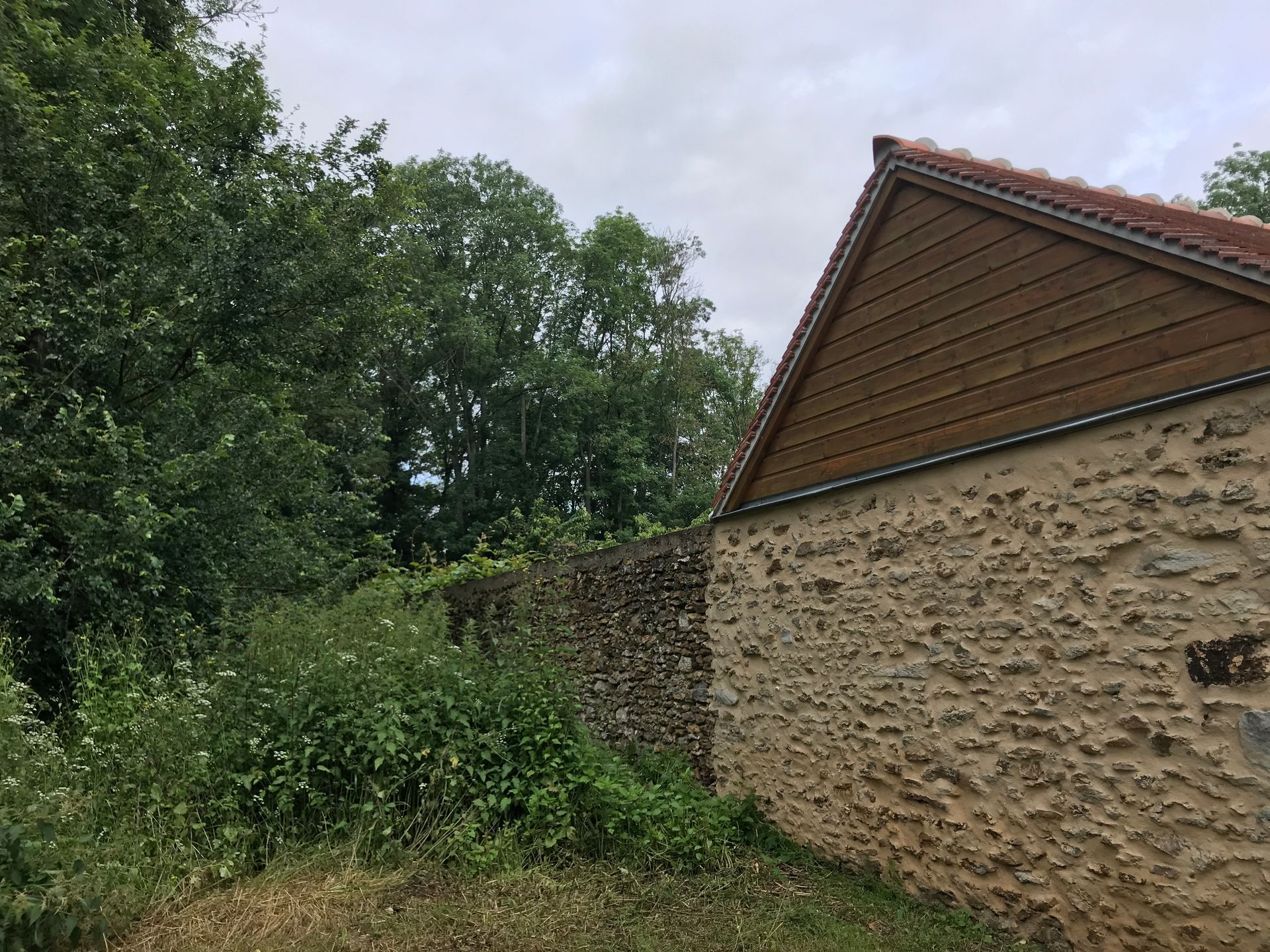 Un mur de pierre jouxte une végétation luxuriante et des arbres, avec le toit d'une maison sous un ciel nuageux.