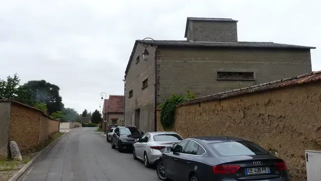 Rue étroite de village avec des voitures garées à côté de vieux bâtiments en pierre sous un ciel couvert