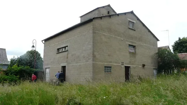 Maison en briques grises au toit en pente, bordée de hautes herbes et de quelques personnes à l'extérieur.