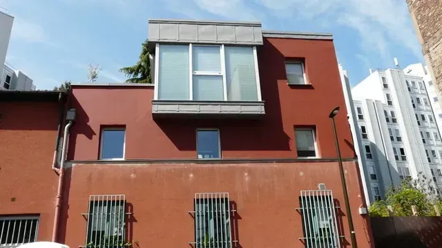 Maison en stuc rouge avec une grande baie vitrée et des fenêtres basses grillagées sous un ciel bleu