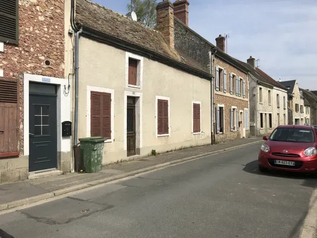 Rue de village tranquille bordée de maisons mitoyennes, une voiture rouge garée et une poubelle verte