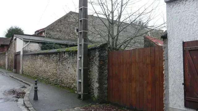 Rue résidentielle pluvieuse avec un mur de pierre, un portail en bois, un poteau électrique et des toits de maisons.