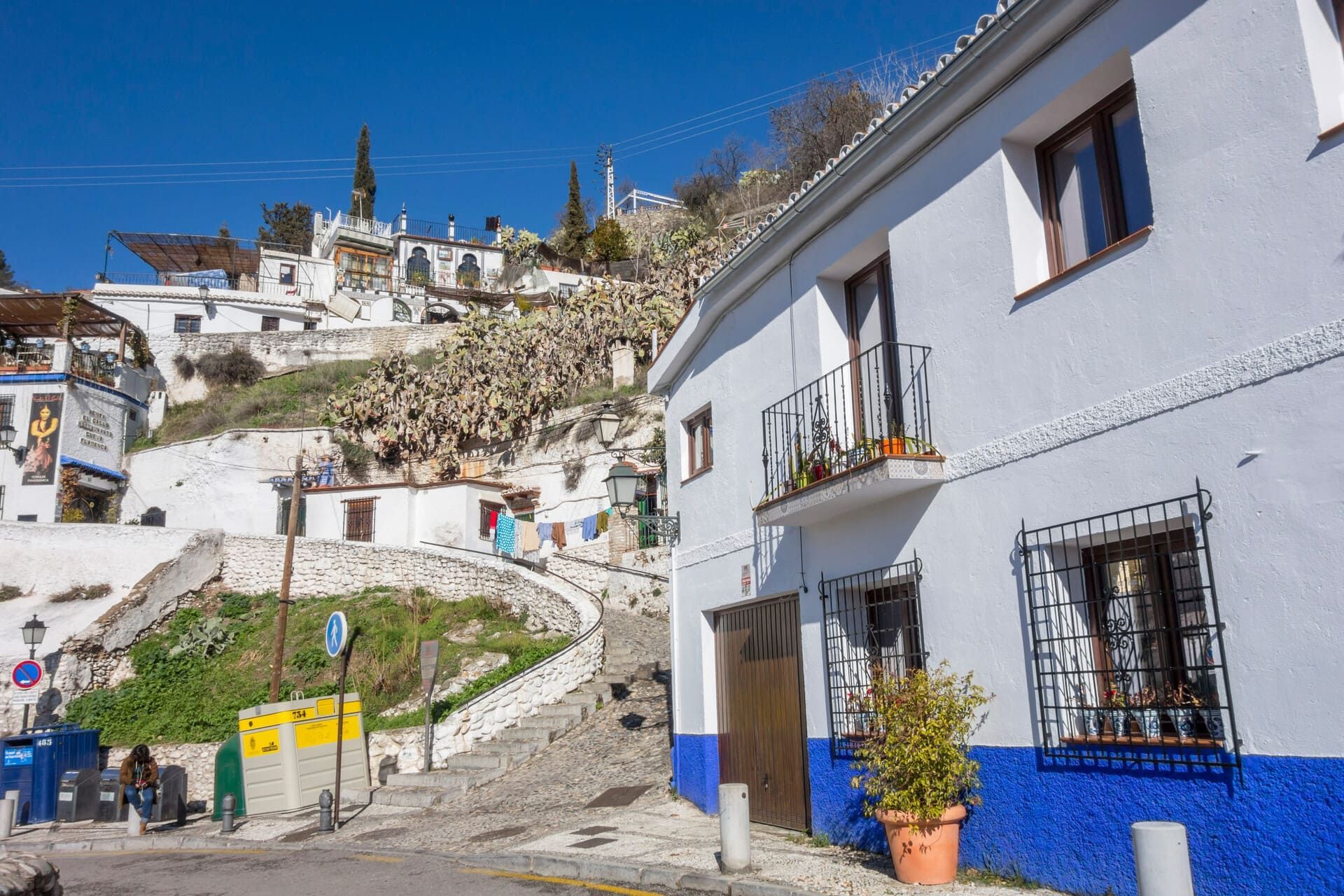 Calle de pueblo en la ladera de una colina, blanca y azul, con casas blancas, camino de piedra y cielo azul claro.