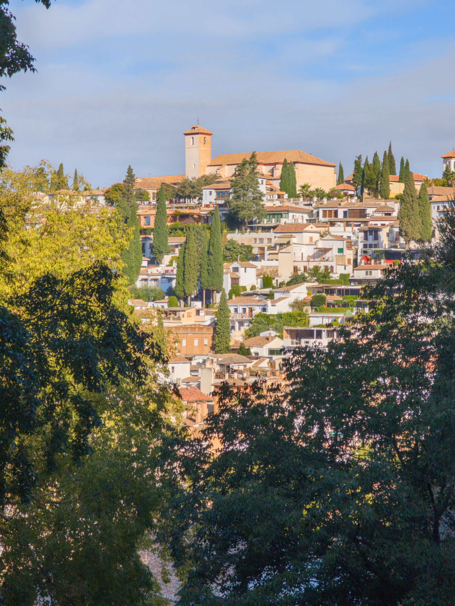 Pueblo soleado en la ladera de una colina con tejados de terracota y una torre de piedra sobre los árboles.
