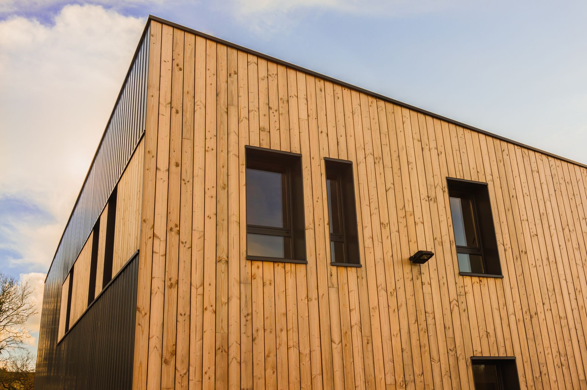 Bâtiment moderne revêtu de bois avec fenêtres sombres, vu d'angle contre un ciel nuageux.