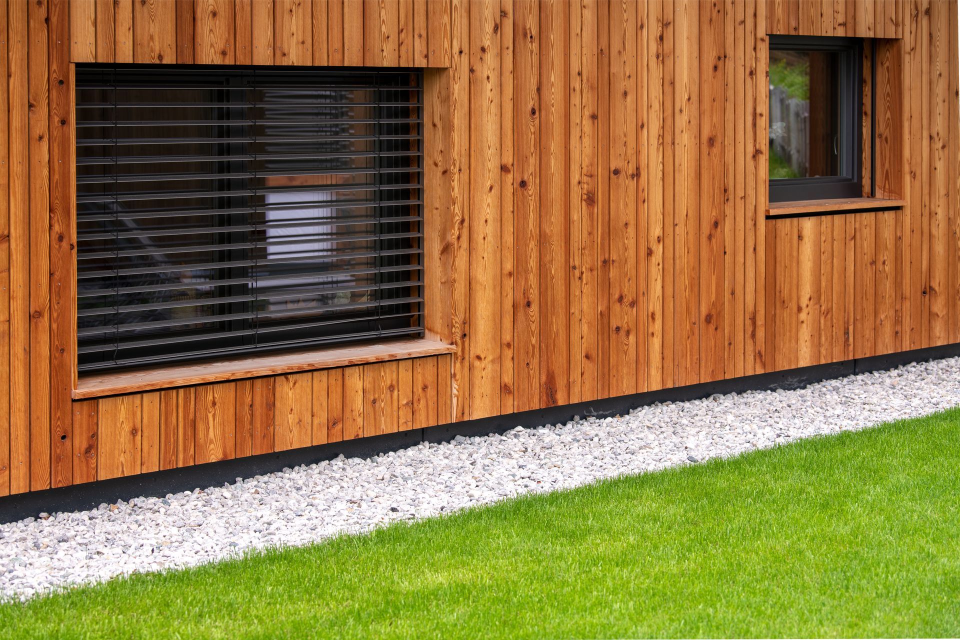 Extérieur d'un bâtiment en bois avec deux fenêtres, entouré de gravier et d'herbe verte.