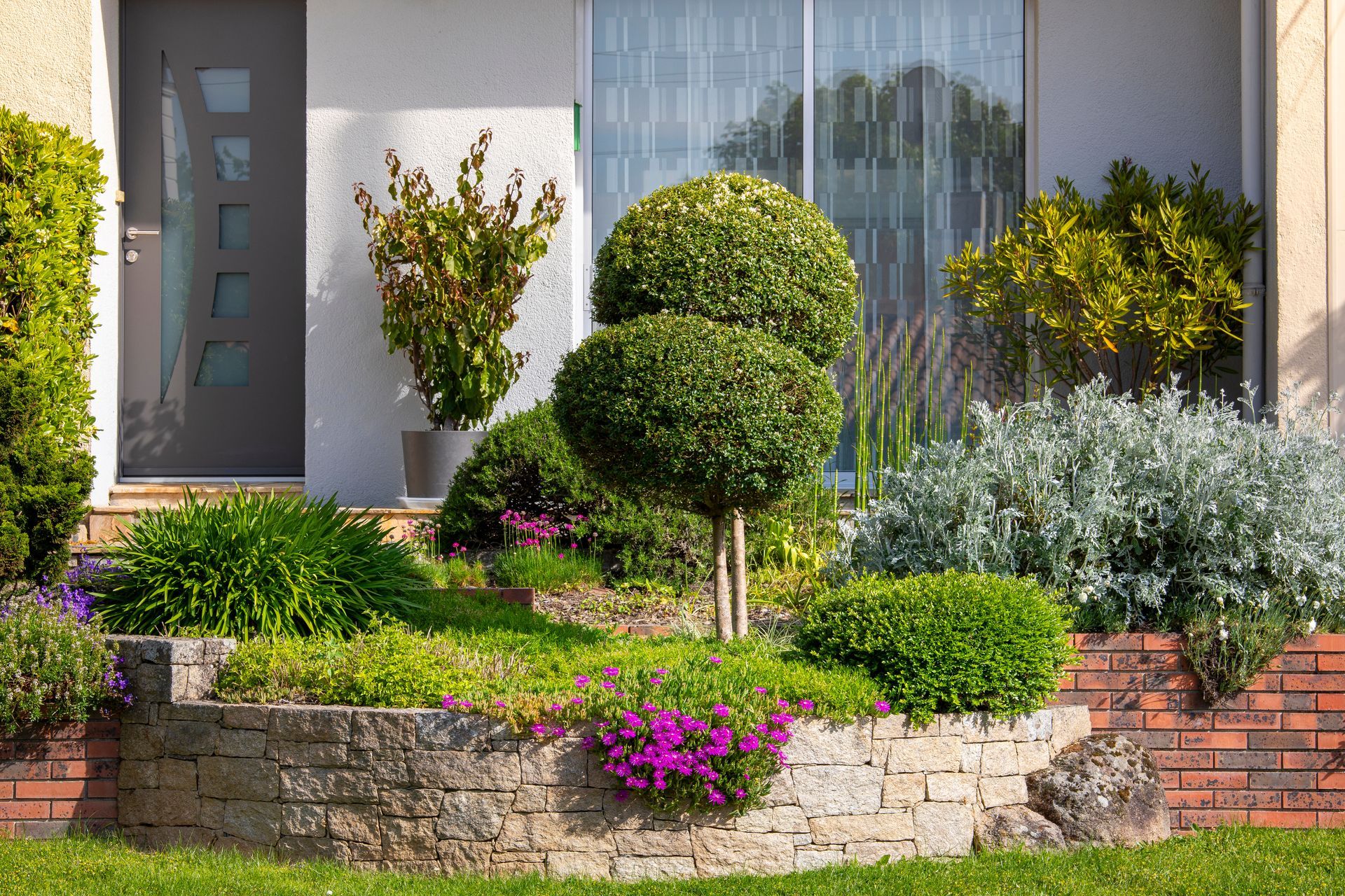 Jardin bien entretenu avec un mur de pierre à plusieurs niveaux, comportant des arbustes verts et des fleurs roses devant une maison.