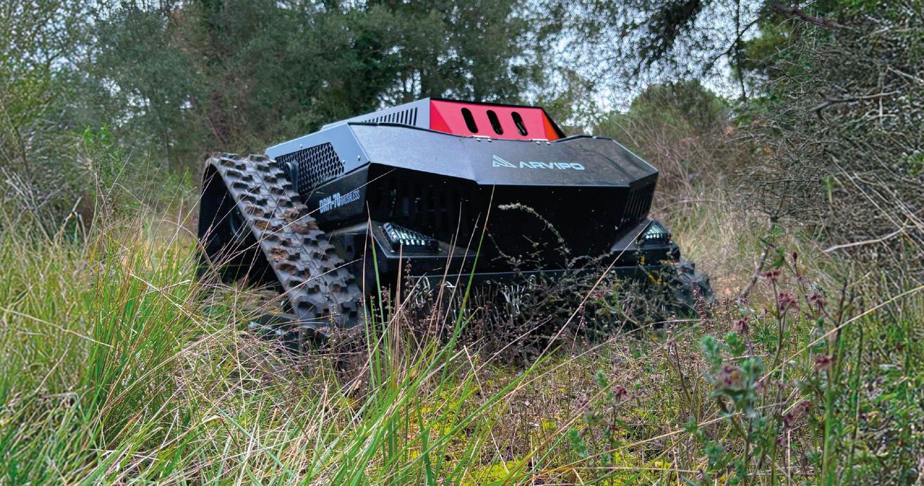 Tondeuse robotisée noir et rouge sur chenilles dans un champ d'herbe, avec des arbres d'automne en arrière-plan.