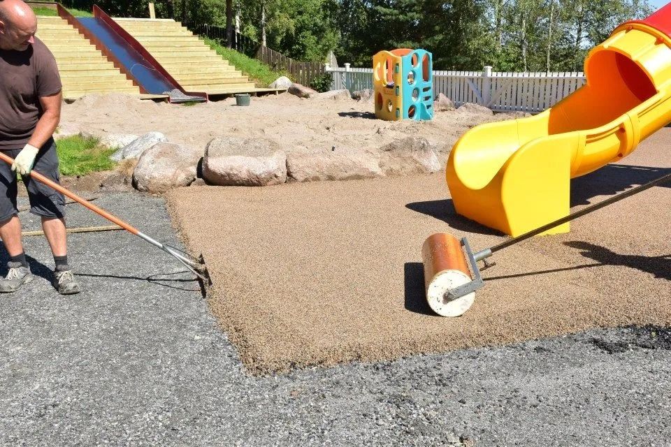 Ein Arbeiter glättet Kies neben einer gelben Spielplatzrutsche und einem Klettergerüst in einem Park.