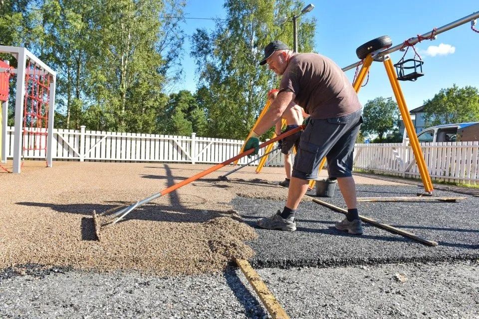 Ein Arbeiter ebnet Kies mit einem langen orangefarbenen Werkzeug auf einer Spielplatzbaustelle.