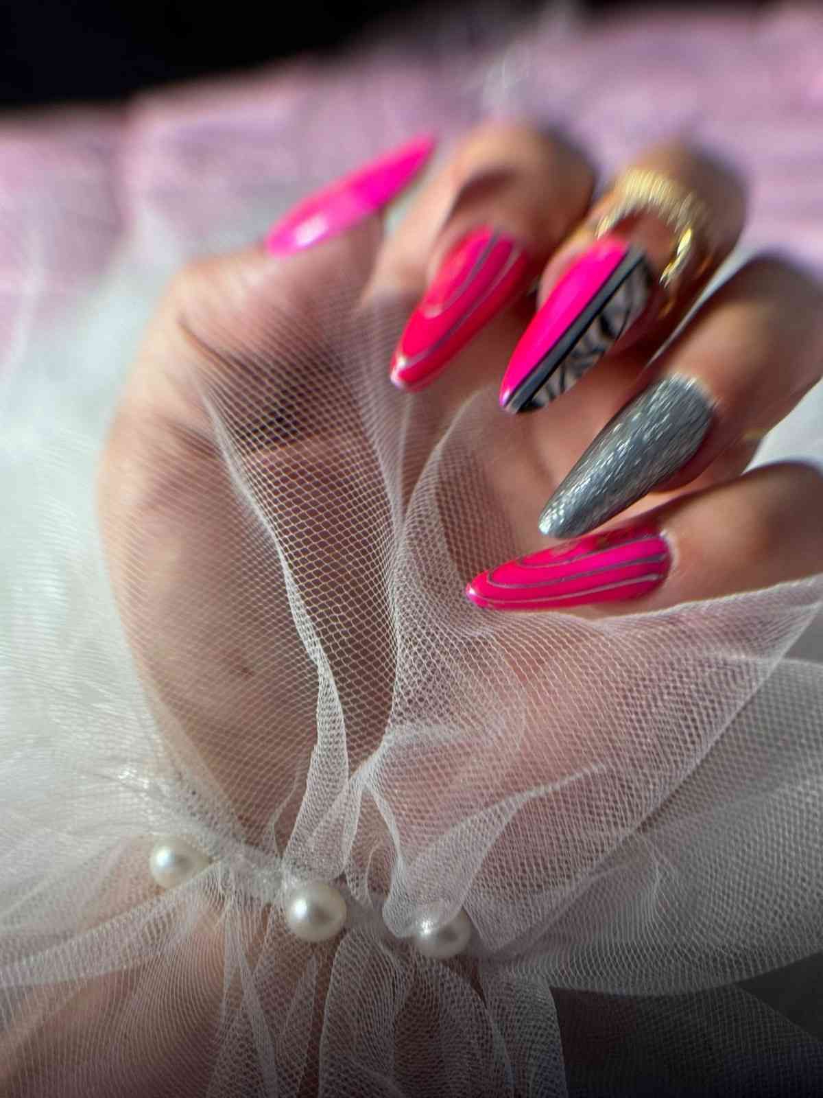 Mano con uñas largas de color rosa neón y plateado estilo stiletto sobre tela blanca transparente con perlas.