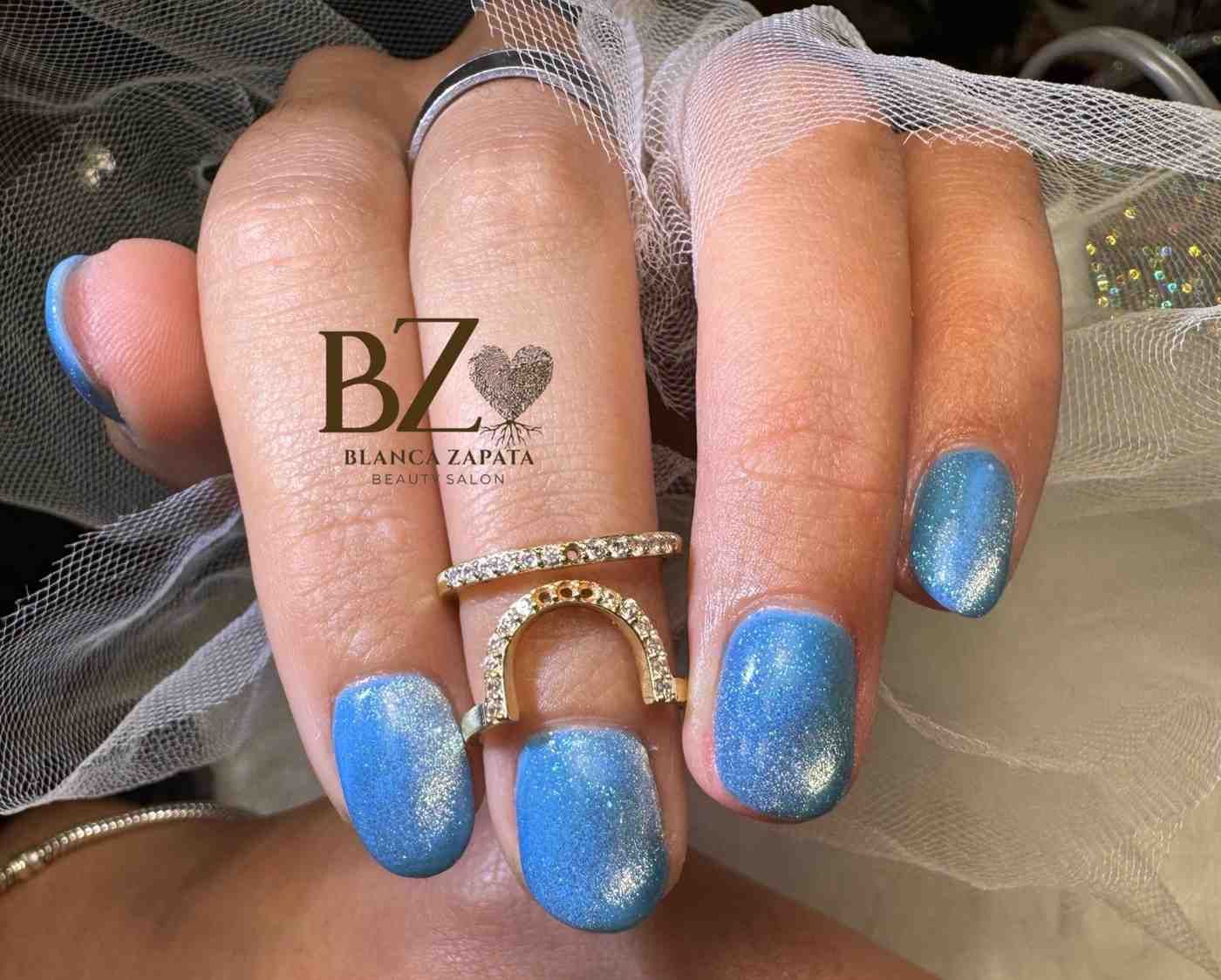Una mano con uñas decoradas con brillantes de color azul y anillos de diamantes, descansa sobre una tela de tul transparente.