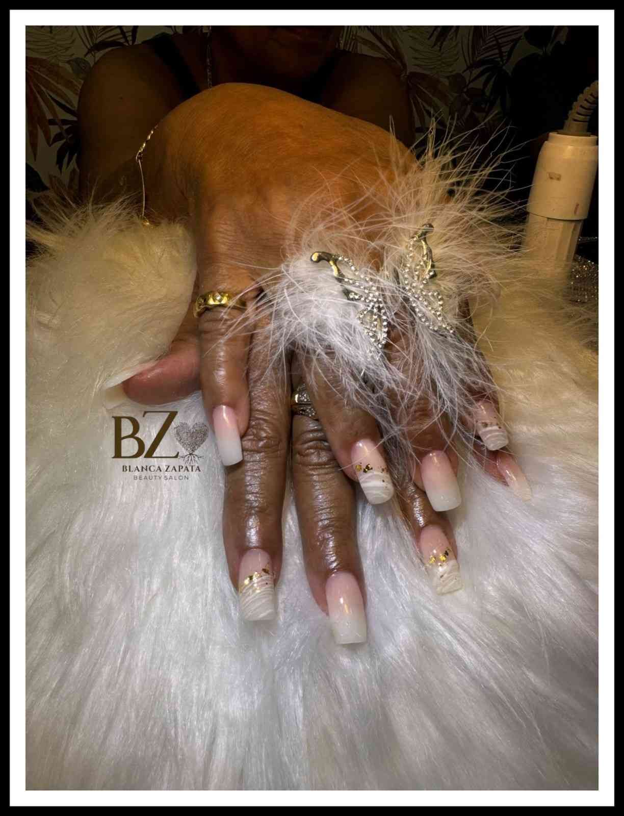 Mano bien cuidada con uñas largas color nude, joyas de pedrería y un accesorio de plumas blancas sobre un fondo de piel.