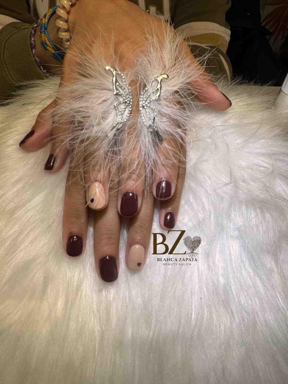 Manos bien cuidadas con uñas de color burdeos oscuro y detalles de plumas enjoyadas que descansan sobre piel blanca.