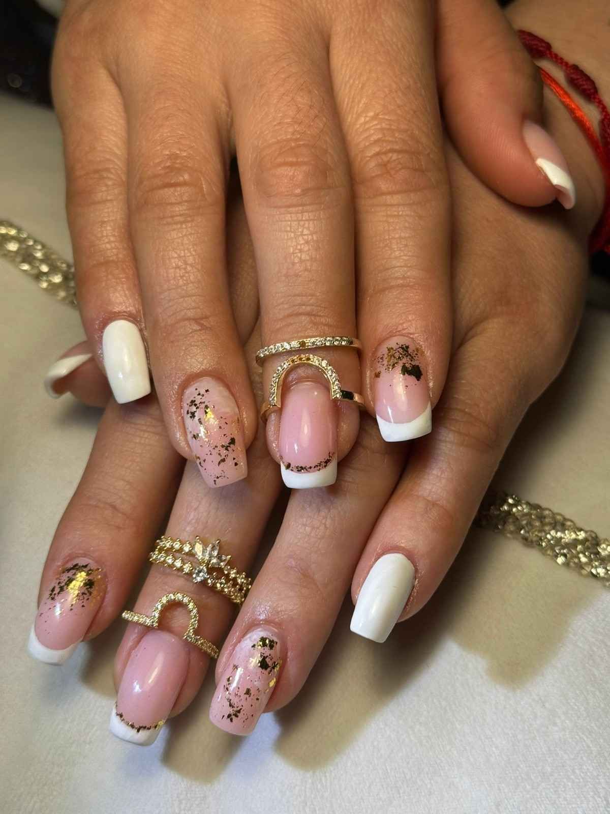 Manos bien cuidadas con uñas blancas y rosas brillantes, luciendo anillos dorados sobre un fondo claro.