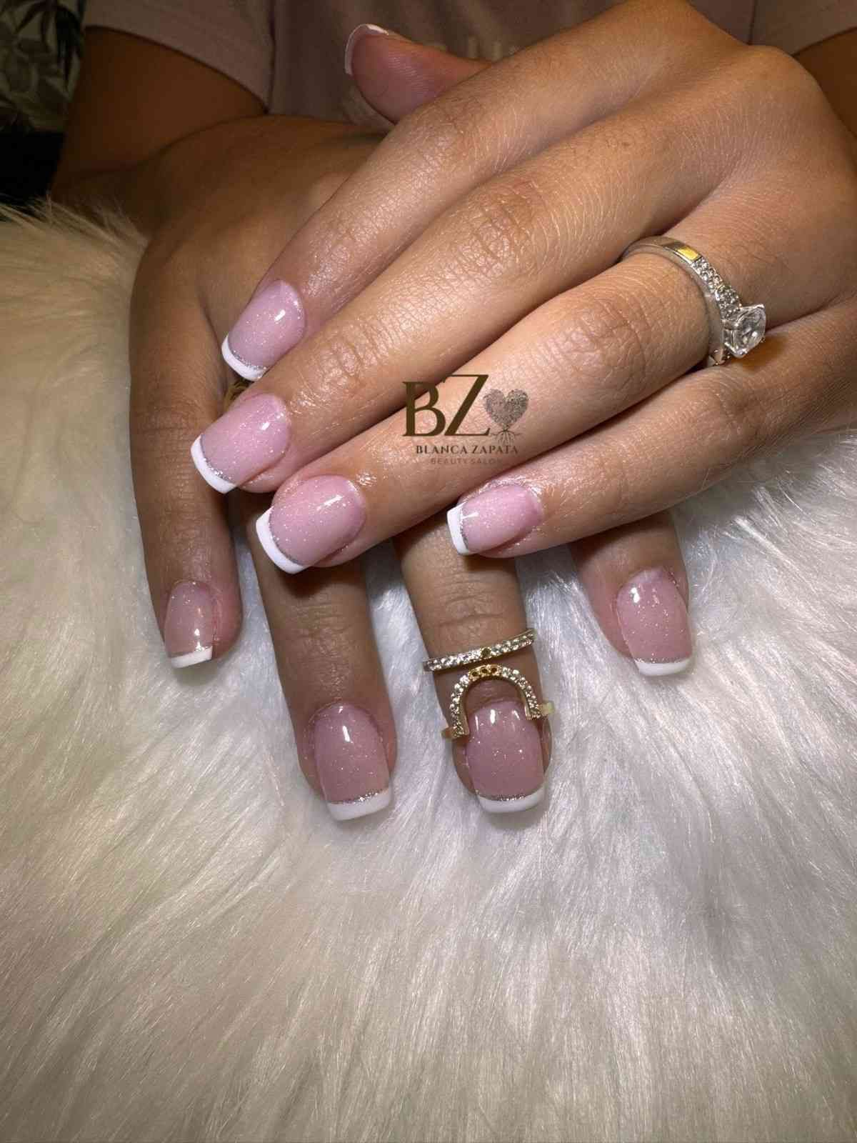 Manos bien cuidadas con uñas rosas y blancas descansando sobre una suave piel blanca, luciendo anillos de oro.