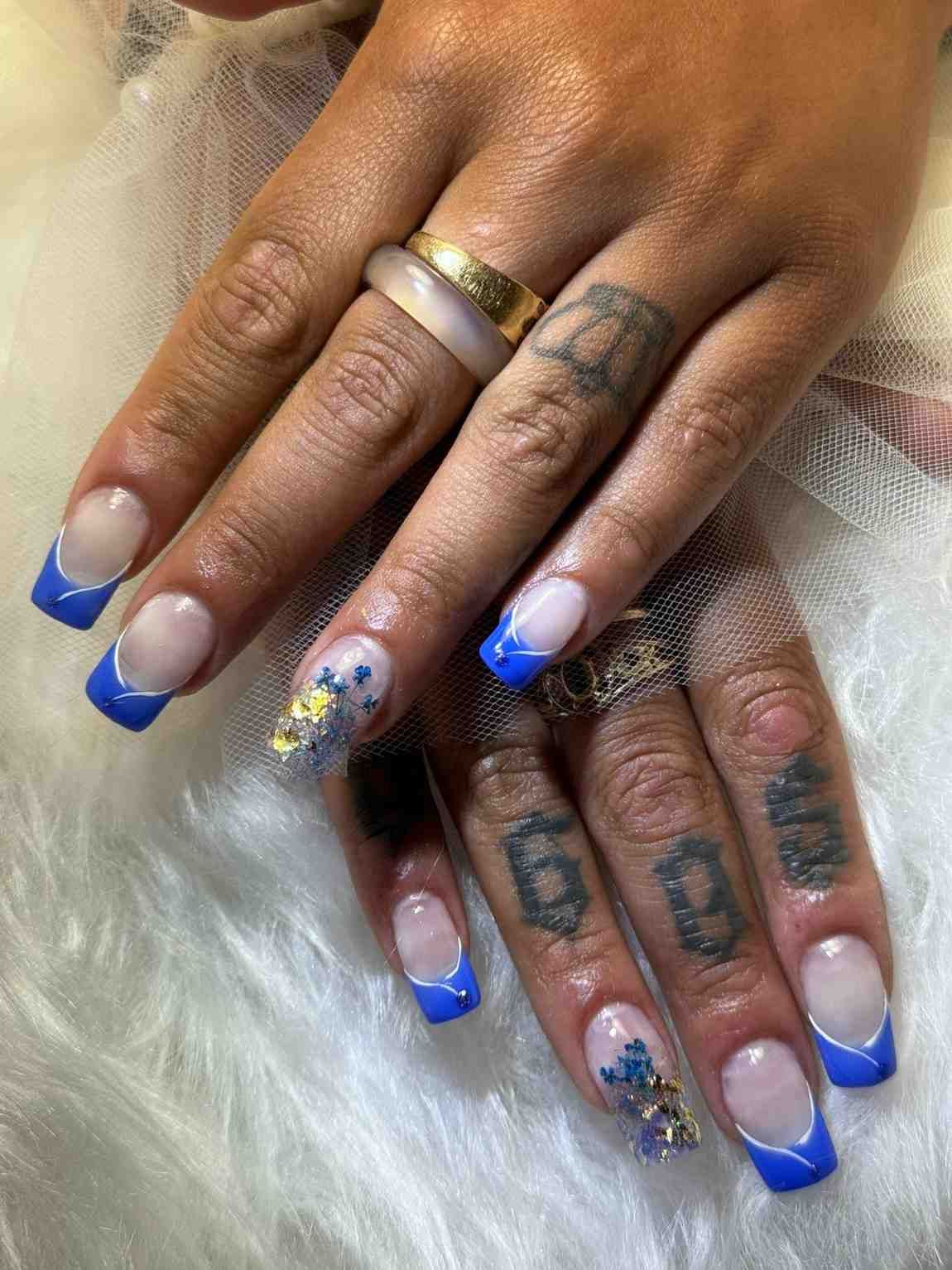 Manos con uñas azules con punta francesa y pequeños diseños florales sobre un fondo de piel blanca.