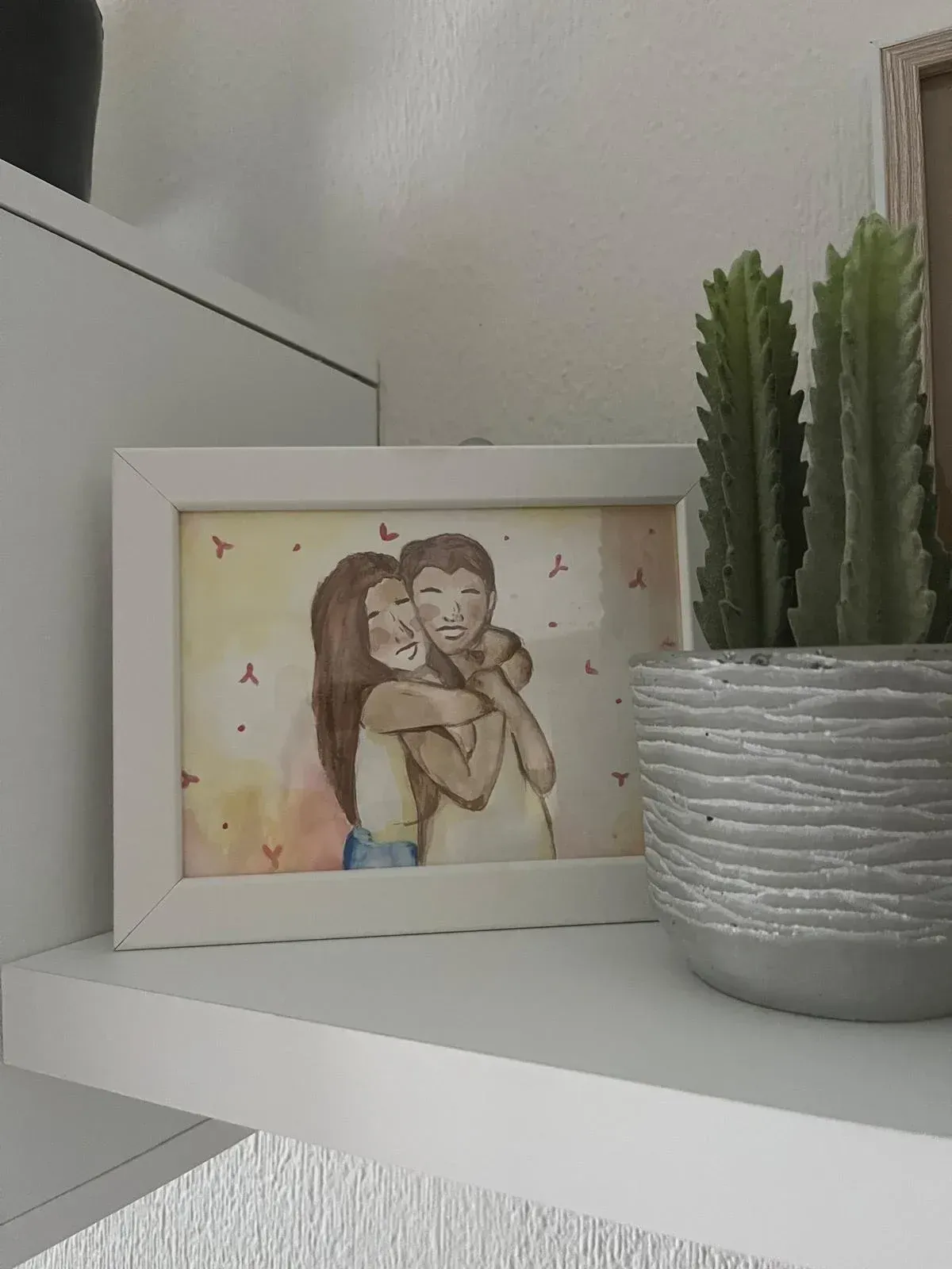 Ilustración enmarcada de una pareja abrazándose sobre una estantería blanca junto a un cactus en maceta.