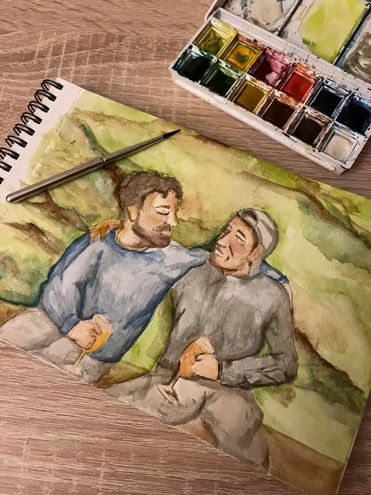 Boceto en acuarela de dos personas sentadas juntas sobre la hierba, con una paleta de pintura y un pincel cerca.