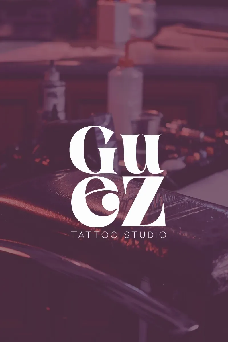 Logotipo de Guaz Tattoo Studio en blanco sobre una estación de trabajo de tatuajes borrosa en tono púrpura.