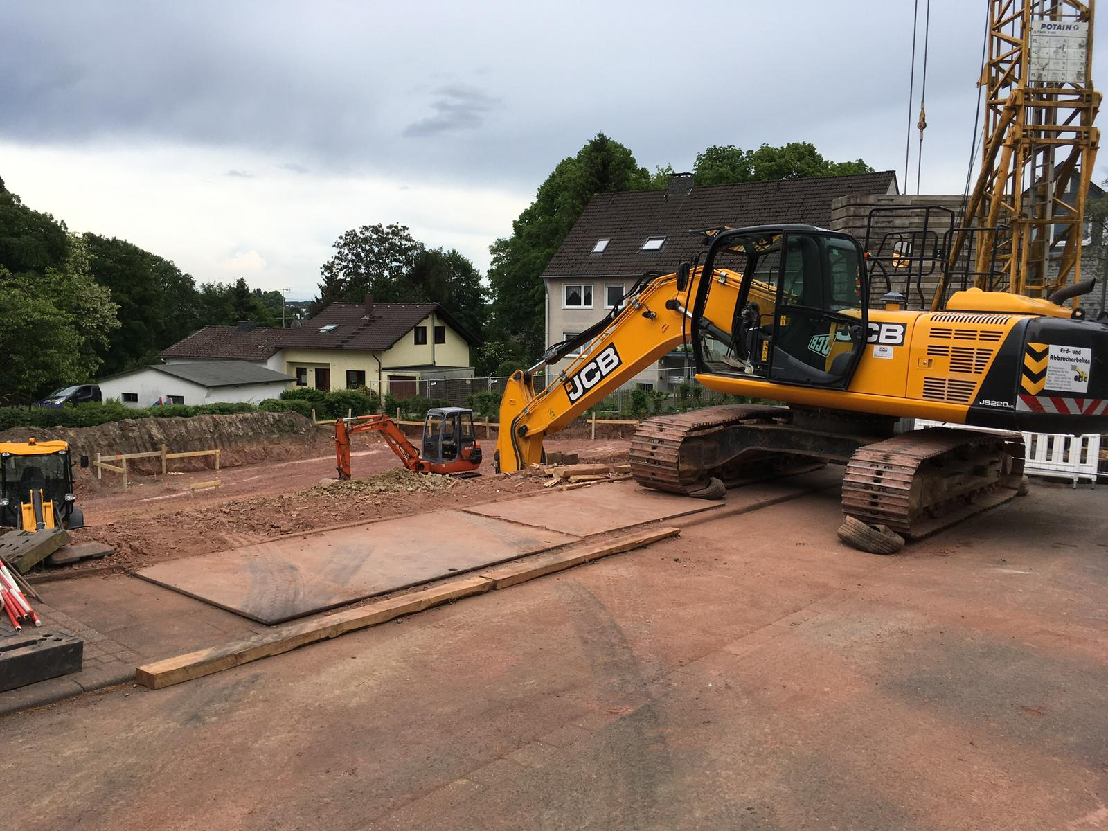 Großer JCB-Kettenbagger auf einer Baustelle mit Stahlplatten vor einer Wohnsiedlung.