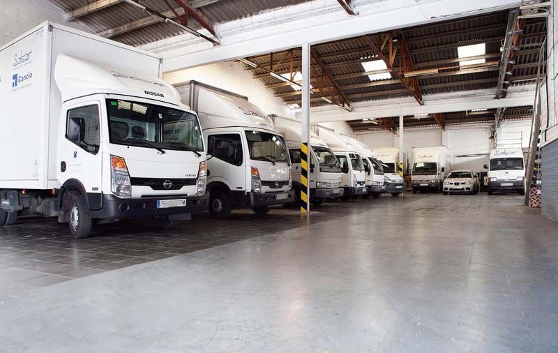 Interior de almacén con varios camiones de reparto blancos estacionados en filas.