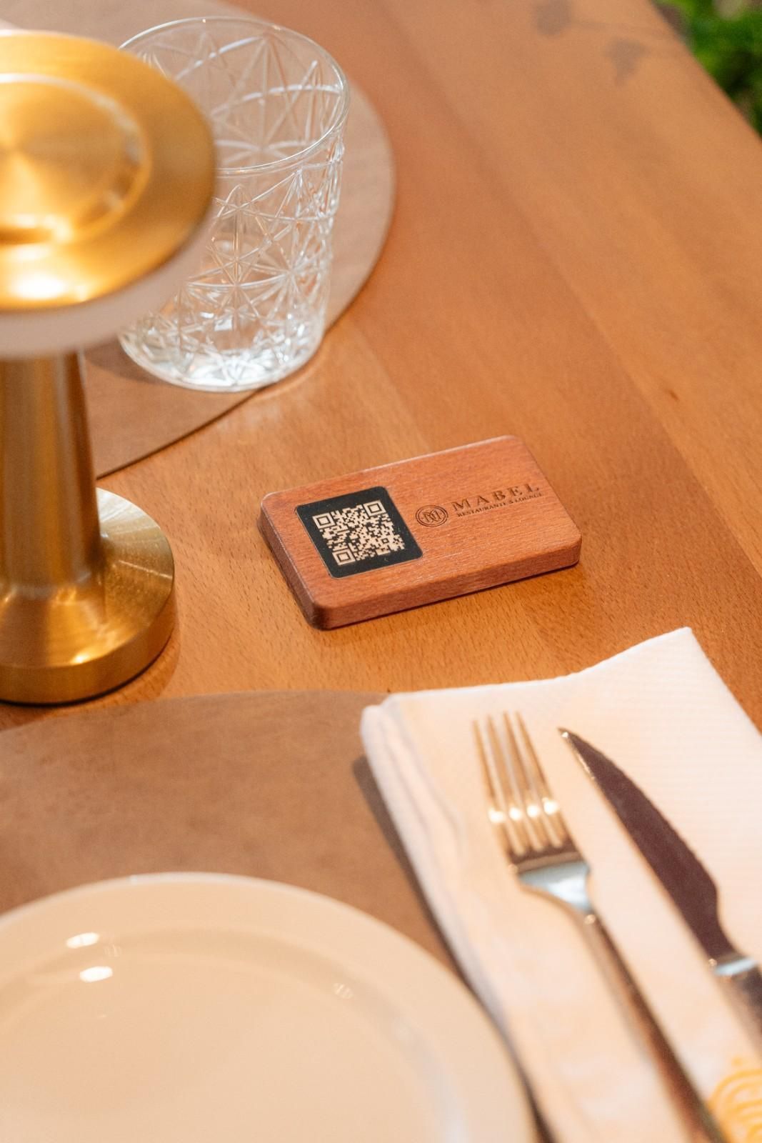 Mesa de madera con una pequeña tarjeta con código QR junto a una lámpara dorada, vaso, servilleta, tenedor y cuchillo.