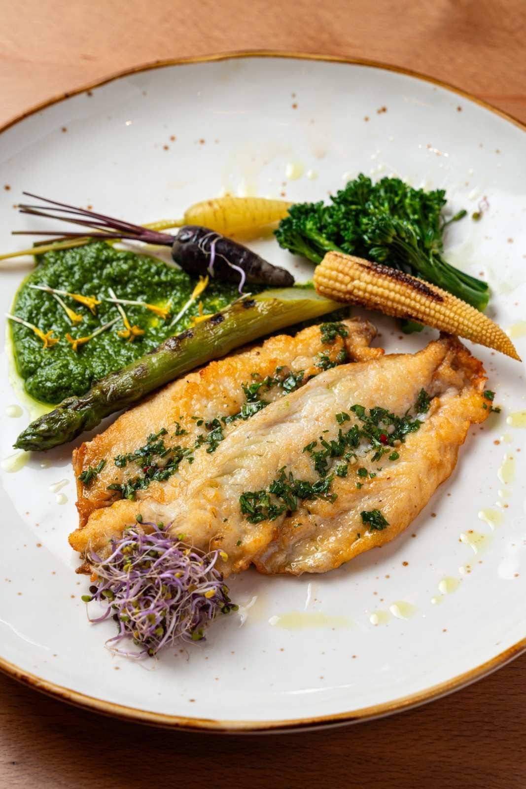 Pescado emplatado con costra de hierbas, espárragos, maíz tierno, verduras y guarnición morada en un plato blanco.