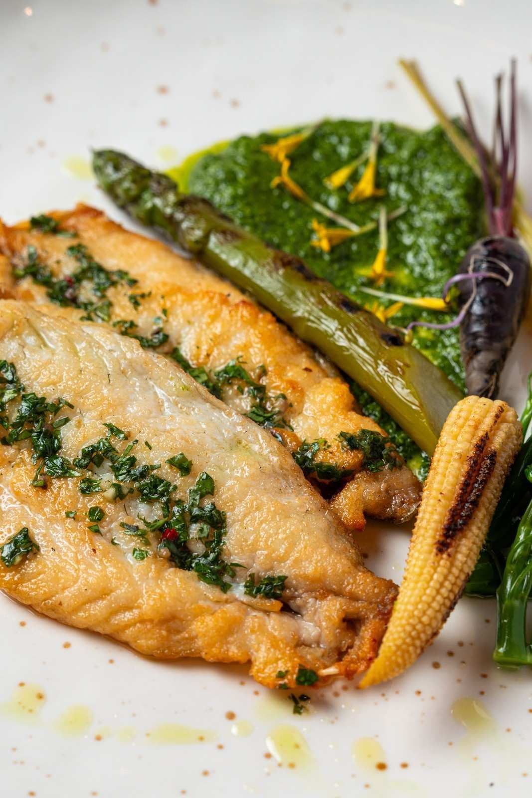Filete de pescado con costra de hierbas, espárragos, maíz tierno, verduras y ralladura de limón en un plato blanco.