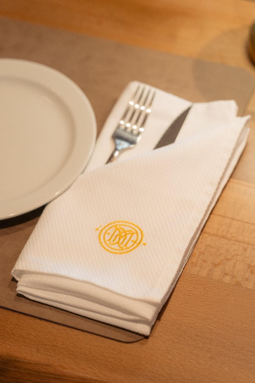 Servilleta blanca doblada con monograma dorado sobre una mesa puesta junto a un plato y cubiertos.
