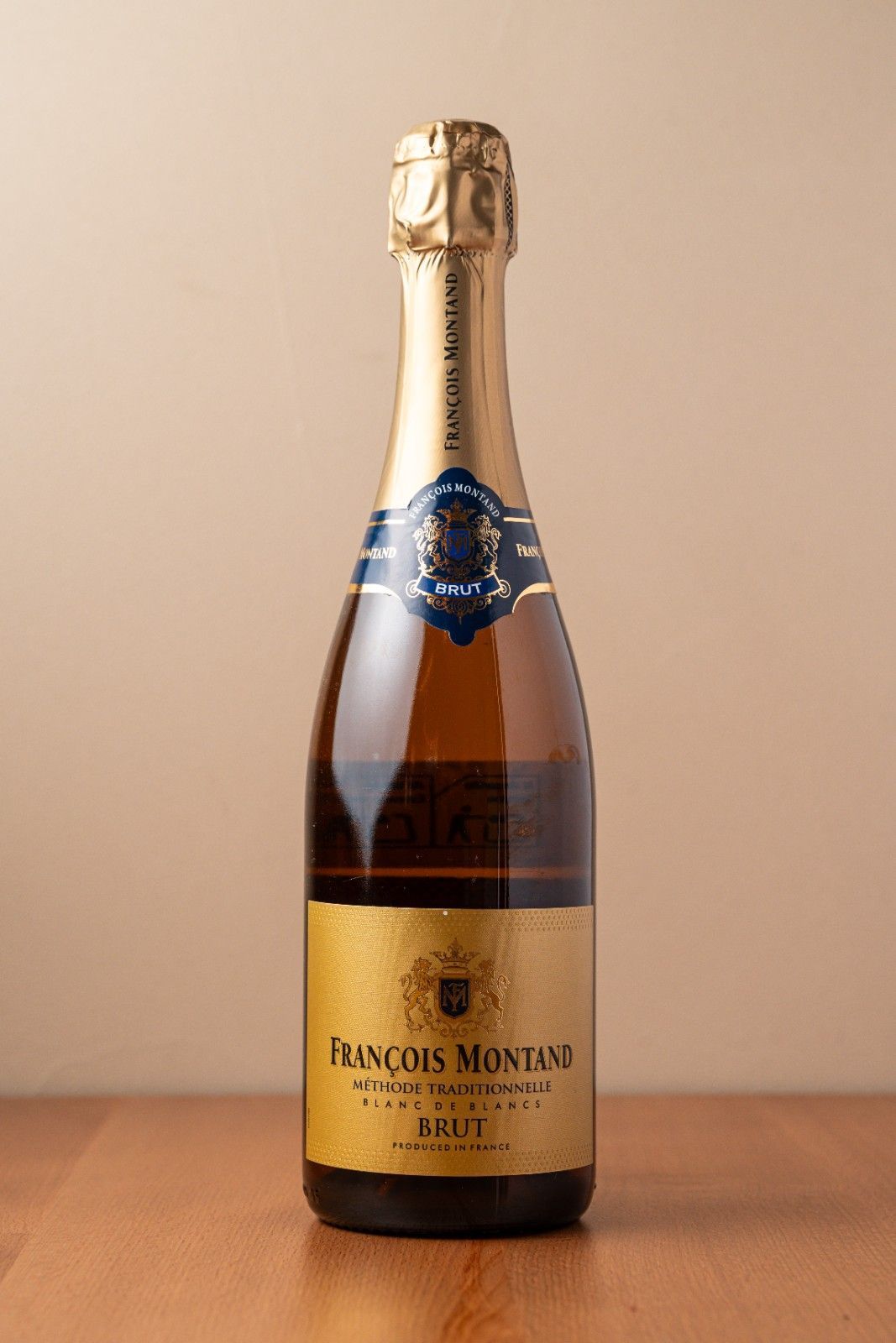 Botella de vino espumoso con lámina de oro y etiqueta azul sobre una mesa de madera con fondo beige.
