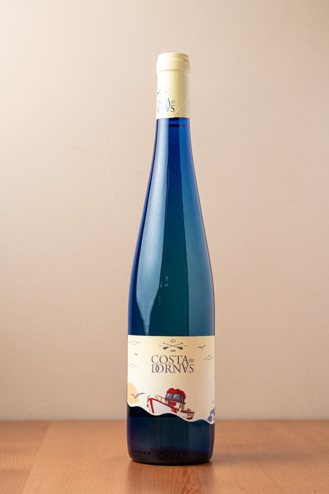 Botella alta de color azul con etiqueta color crema, colocada sobre una mesa de madera con fondo beige.