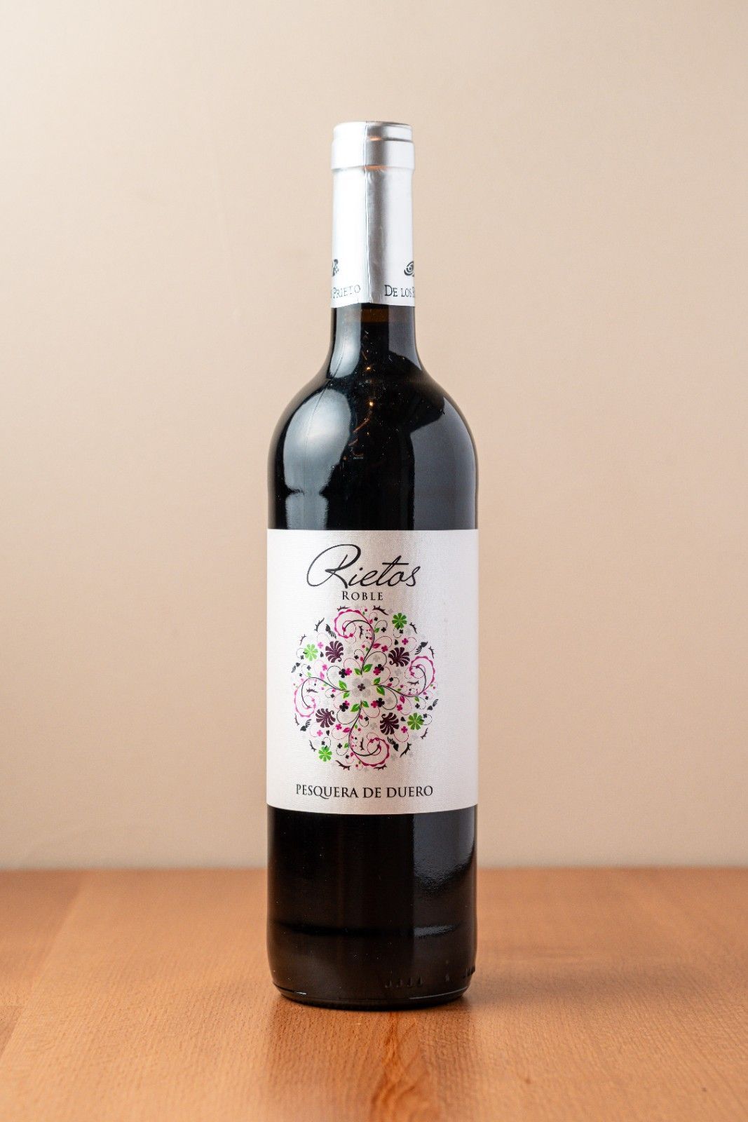 Una botella de vino oscuro con etiqueta floral sobre una mesa de madera con fondo beige.