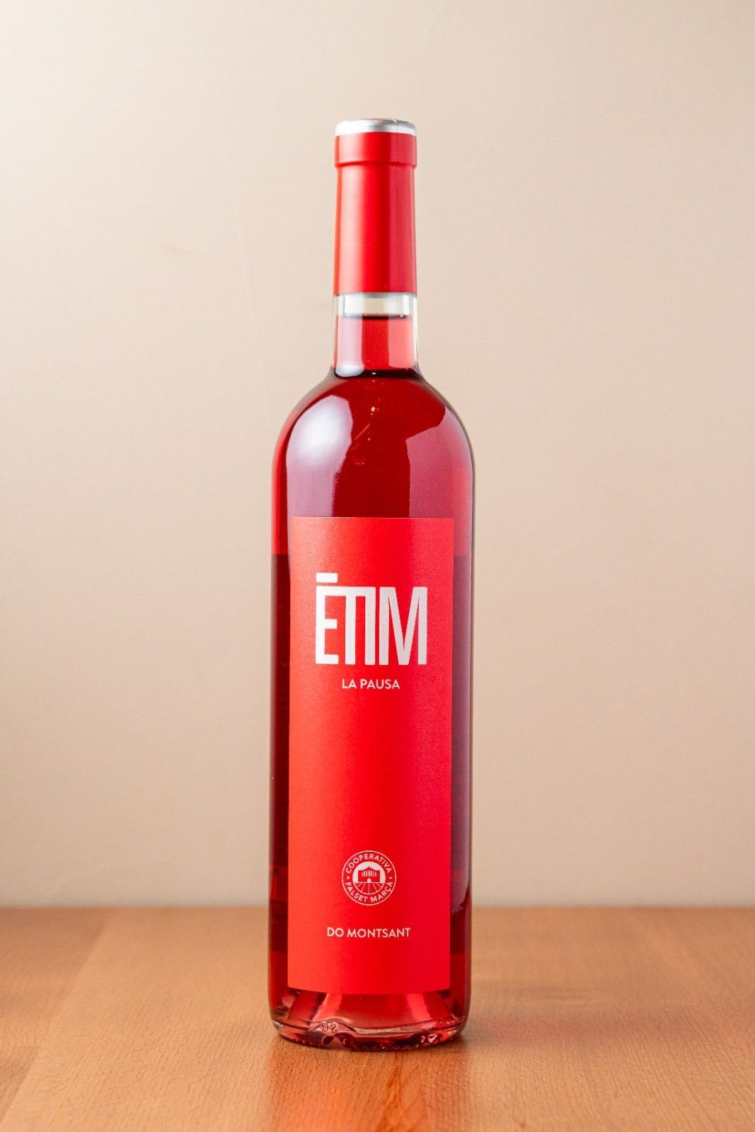 Una botella roja con etiqueta blanca descansa sobre una mesa de madera contra una pared beige.