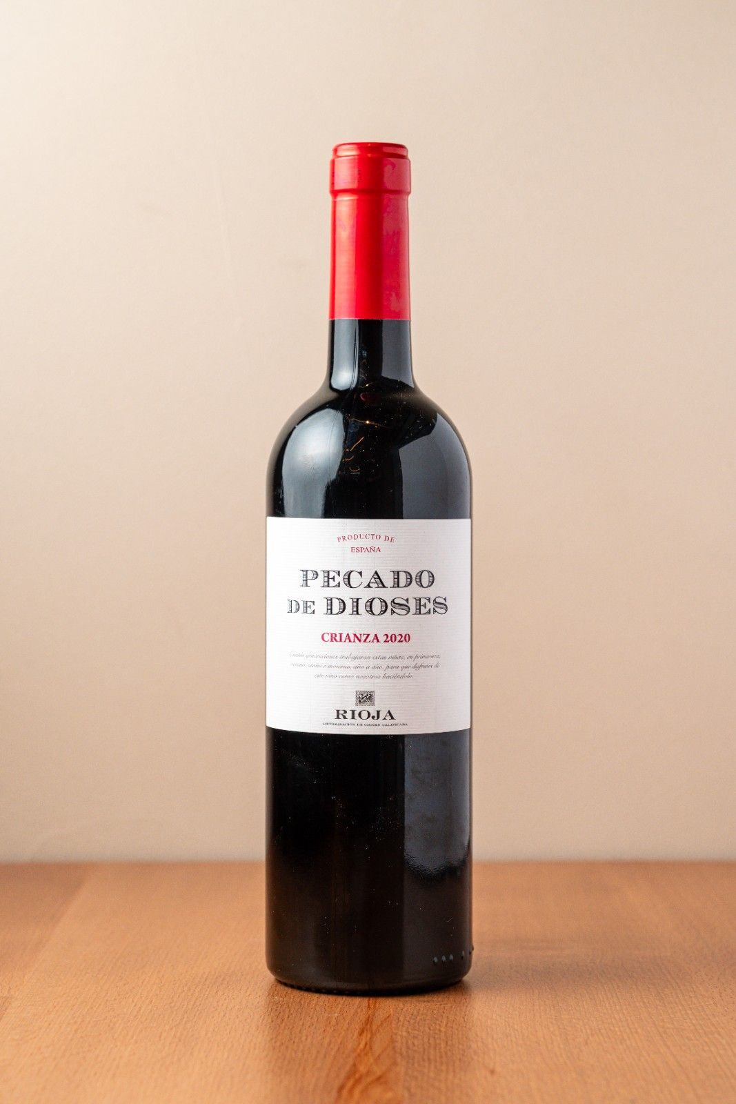 Botella de vino tinto con tapón rojo y etiqueta blanca sobre una mesa de madera contra una pared beige.