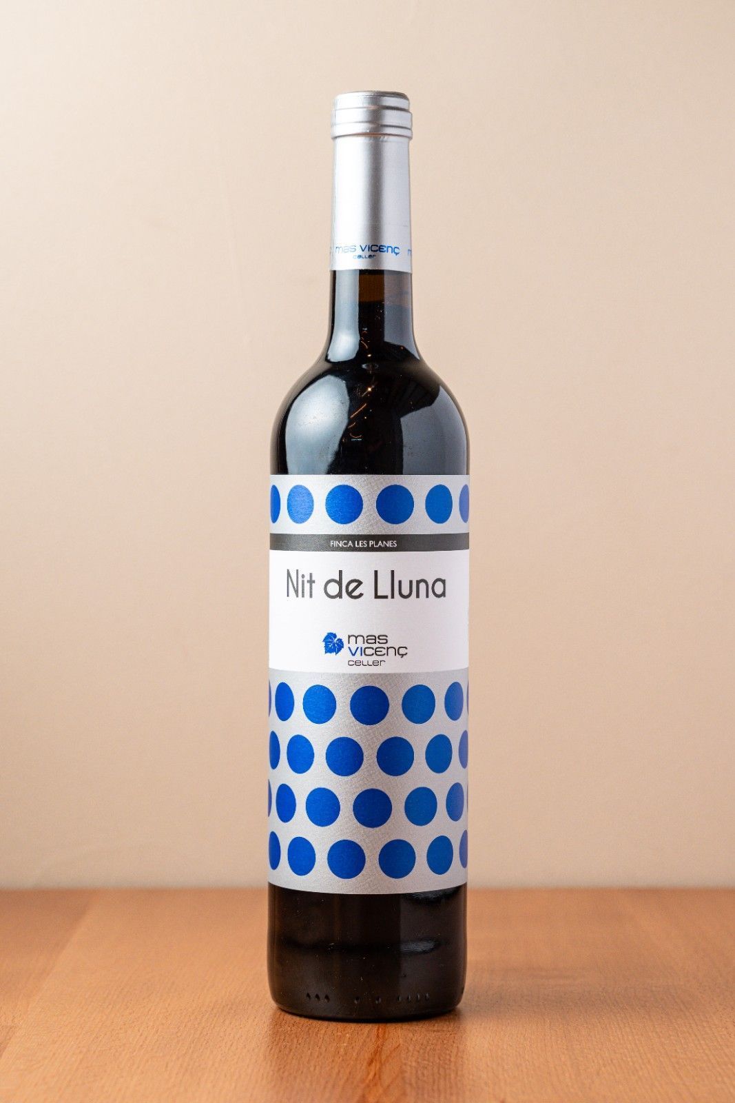 Botella de vino tinto con etiqueta de lunares azules sobre una mesa de madera con fondo beige.