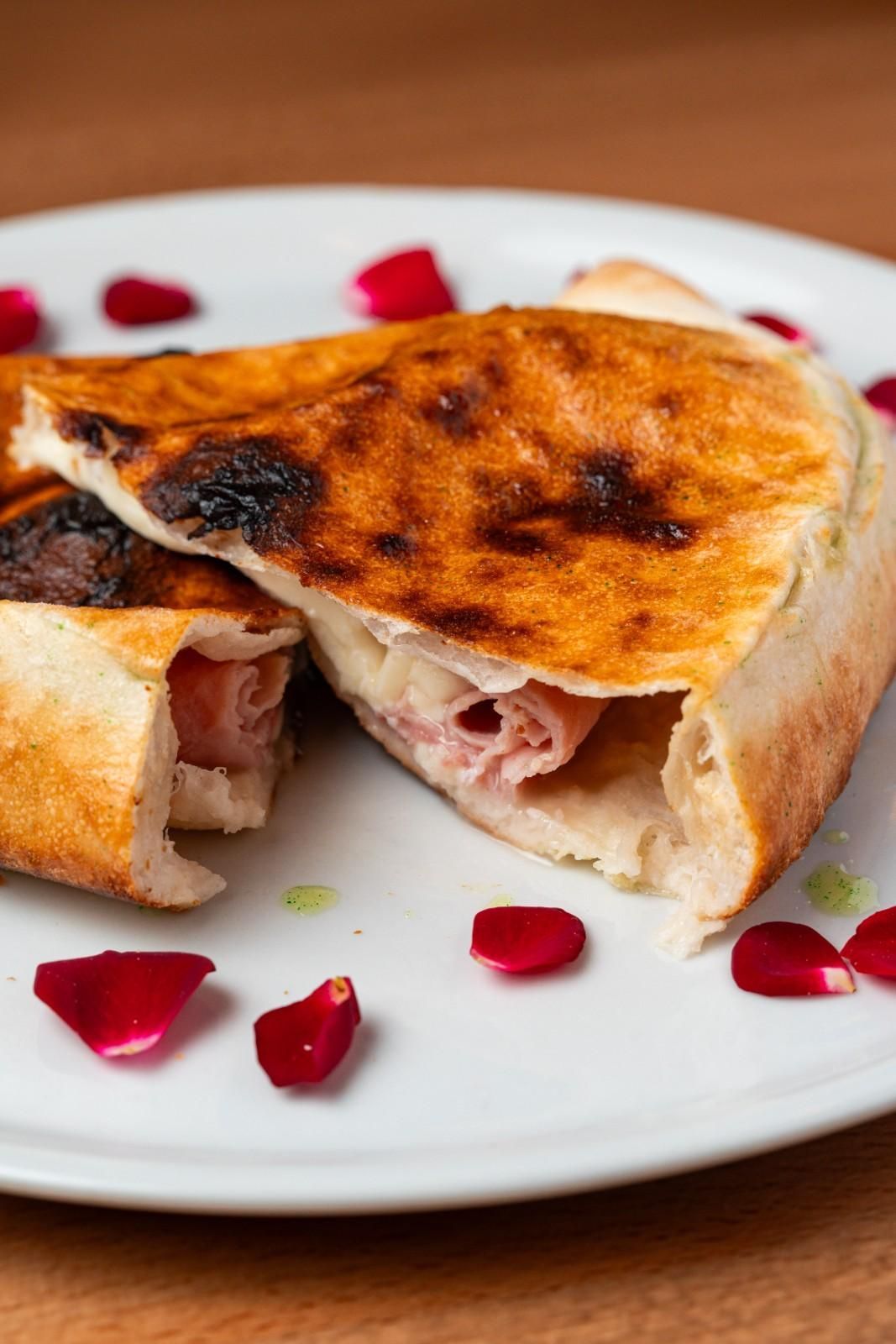 Sándwich de pan plano tostado y doblado con jamón, en un plato blanco con guarnición de pétalos rojos.