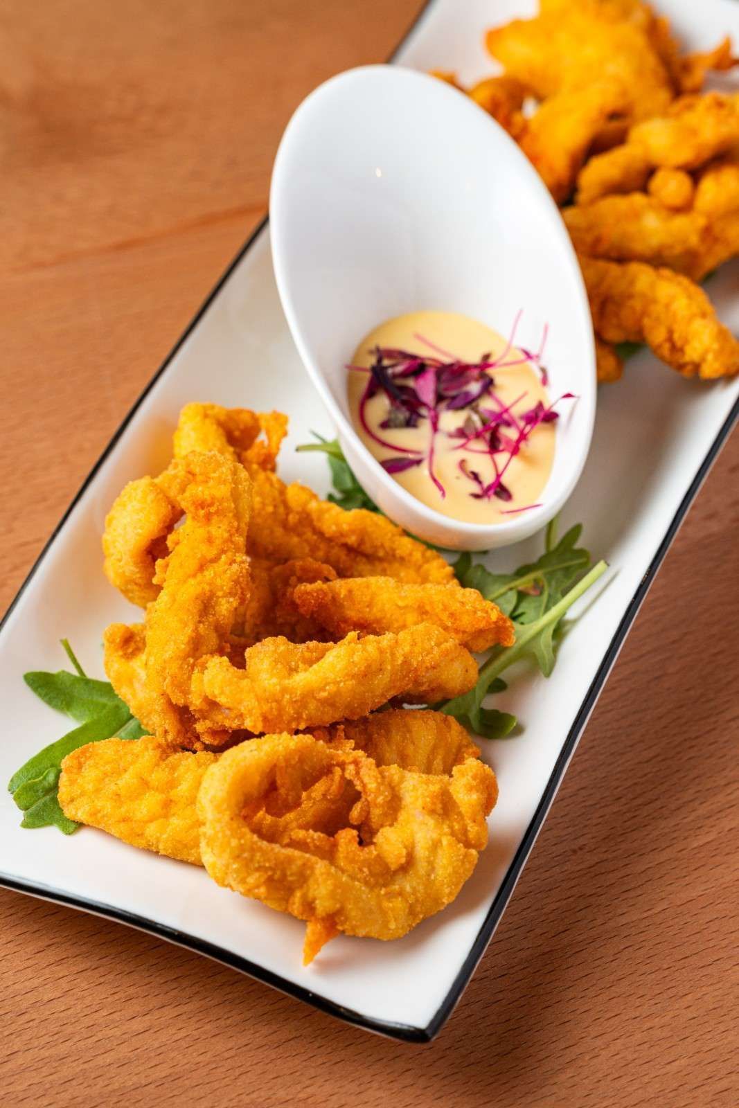 Mariscos fritos dorados emplatados con salsa para mojar y guarnición en un plato rectangular blanco.