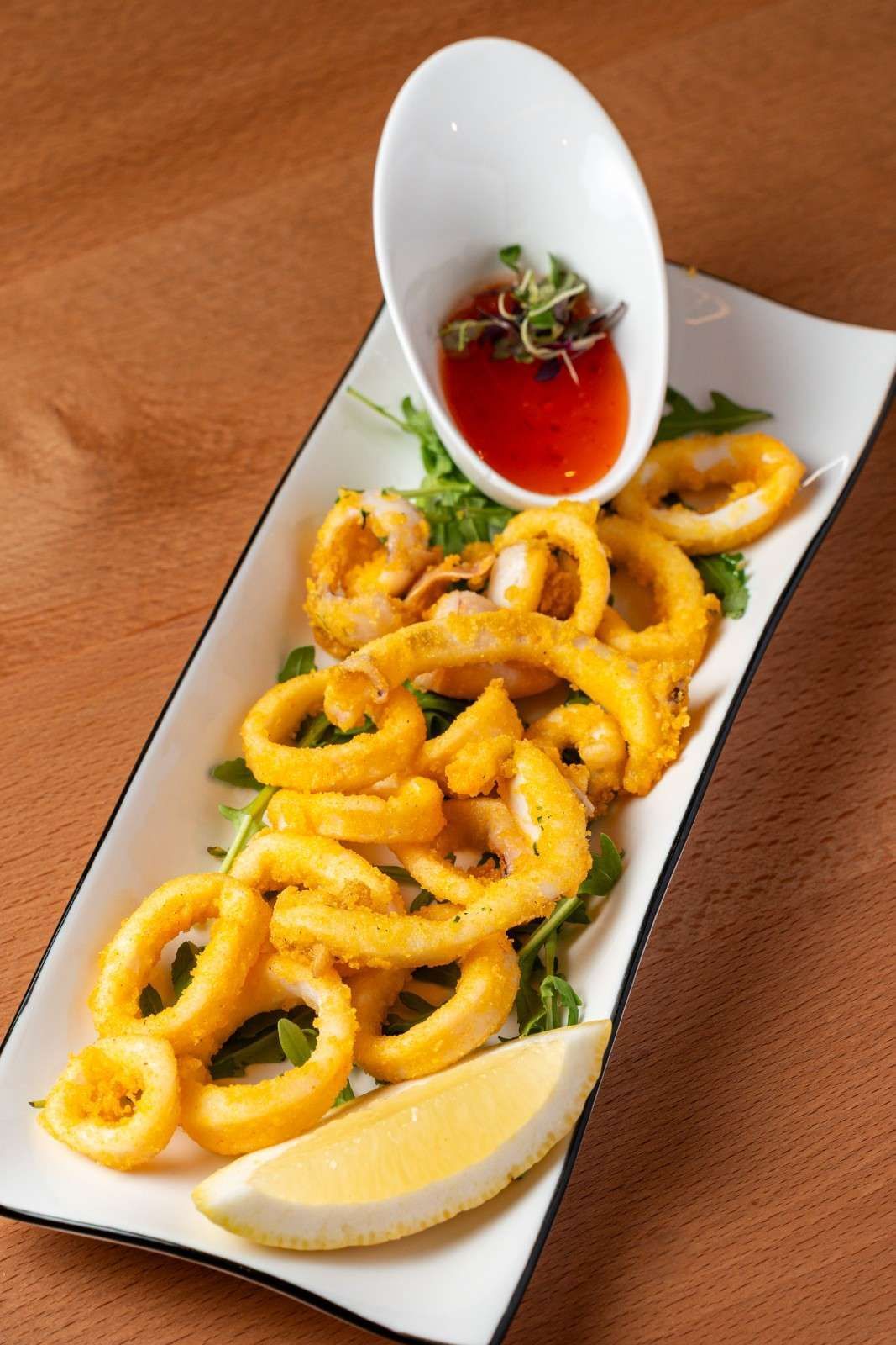 Plato de calamares fritos dorados con rodaja de limón y salsa roja para mojar en una cuchara blanca sobre una mesa de madera.