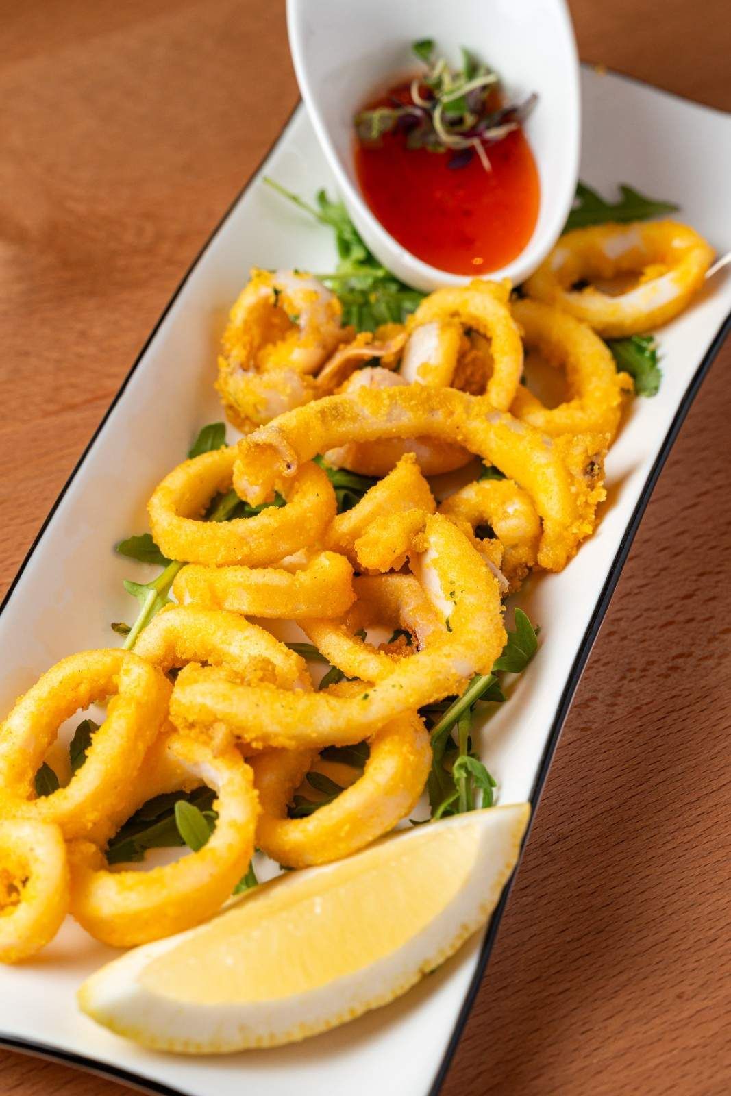 Anillos de calamar fritos en un plato blanco con rodaja de limón y salsa roja para mojar.