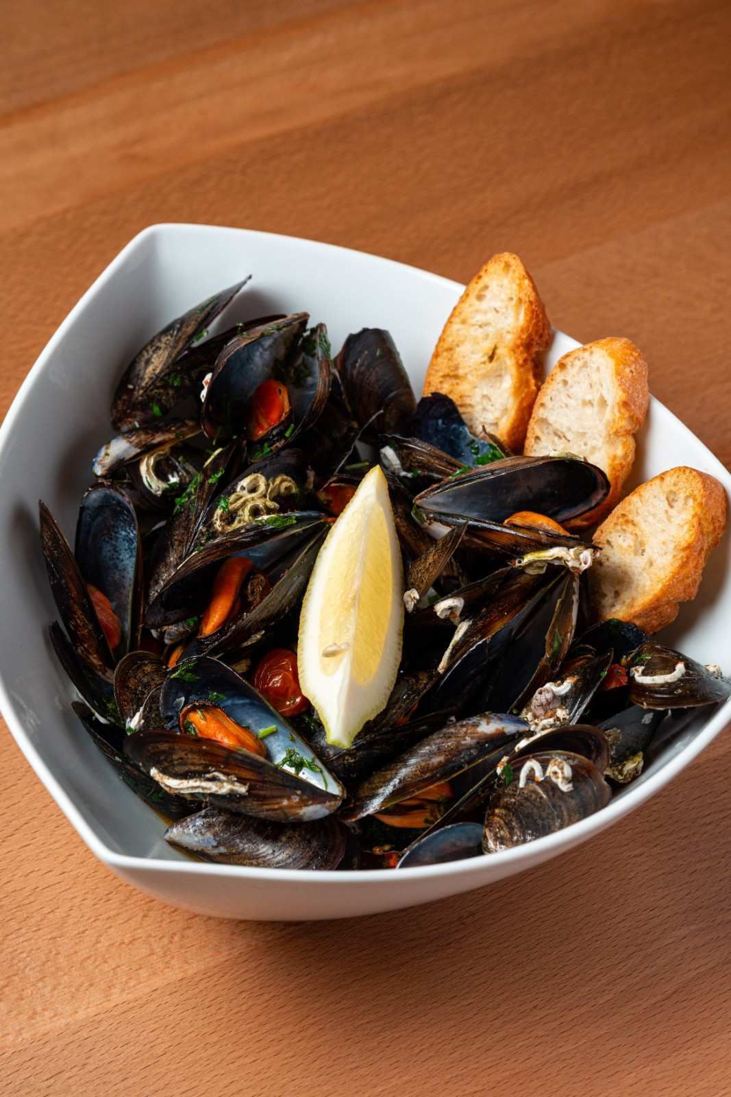 Un bol de mejillones en caldo con una rodaja de limón y pan tostado sobre una mesa de madera.