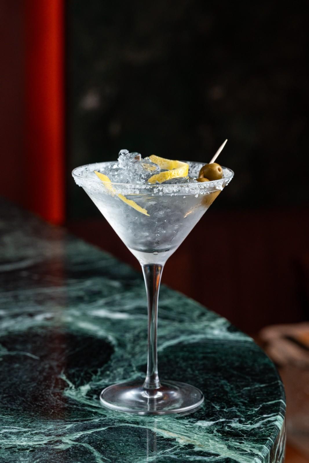Copa de martini con un cóctel transparente, una rodaja de limón y hielo sobre una mesa de mármol oscuro.