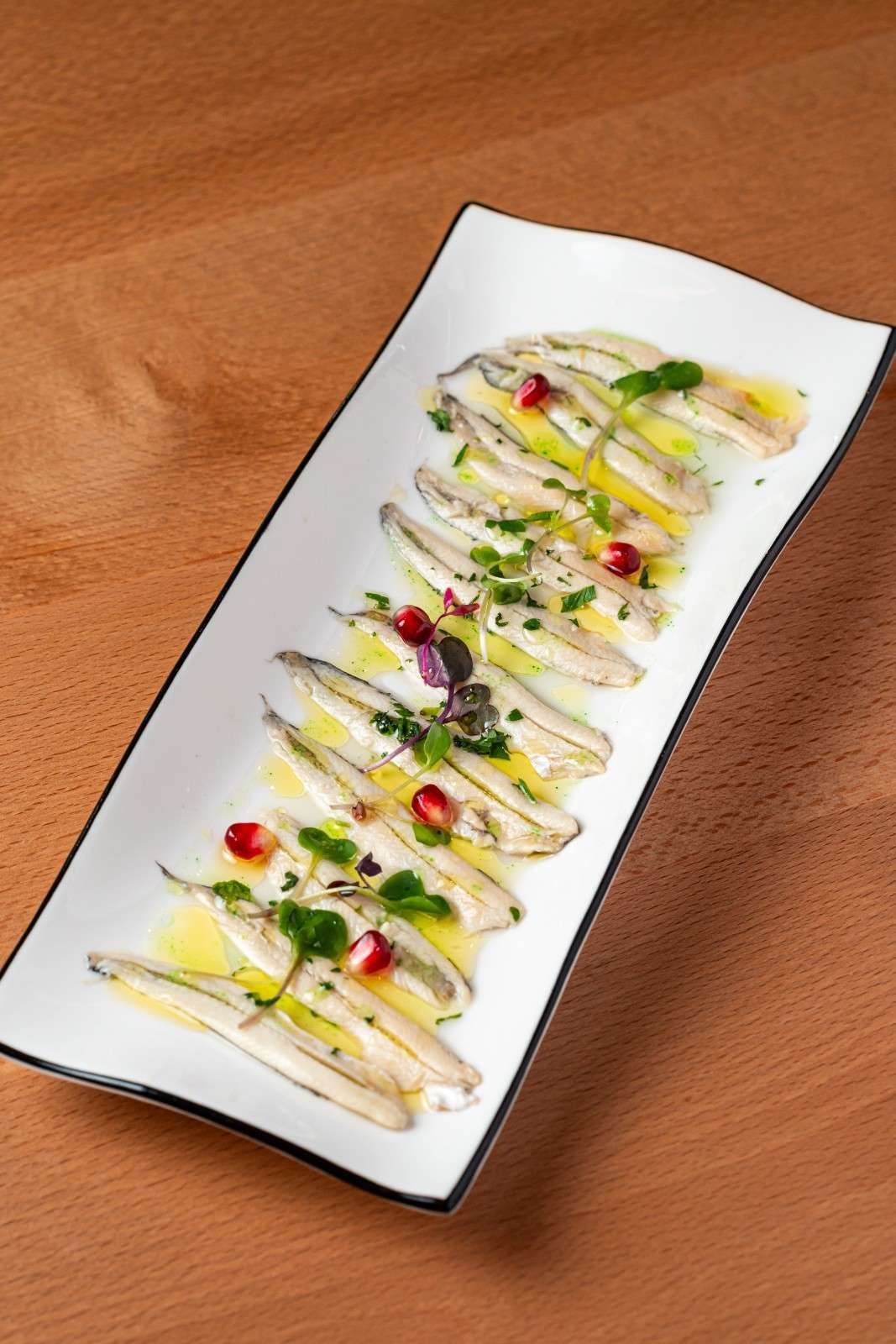 Aperitivo emplatado con finas lonchas de comida rociadas con salsa, adornado con hierbas y trocitos rojos en un plato blanco.