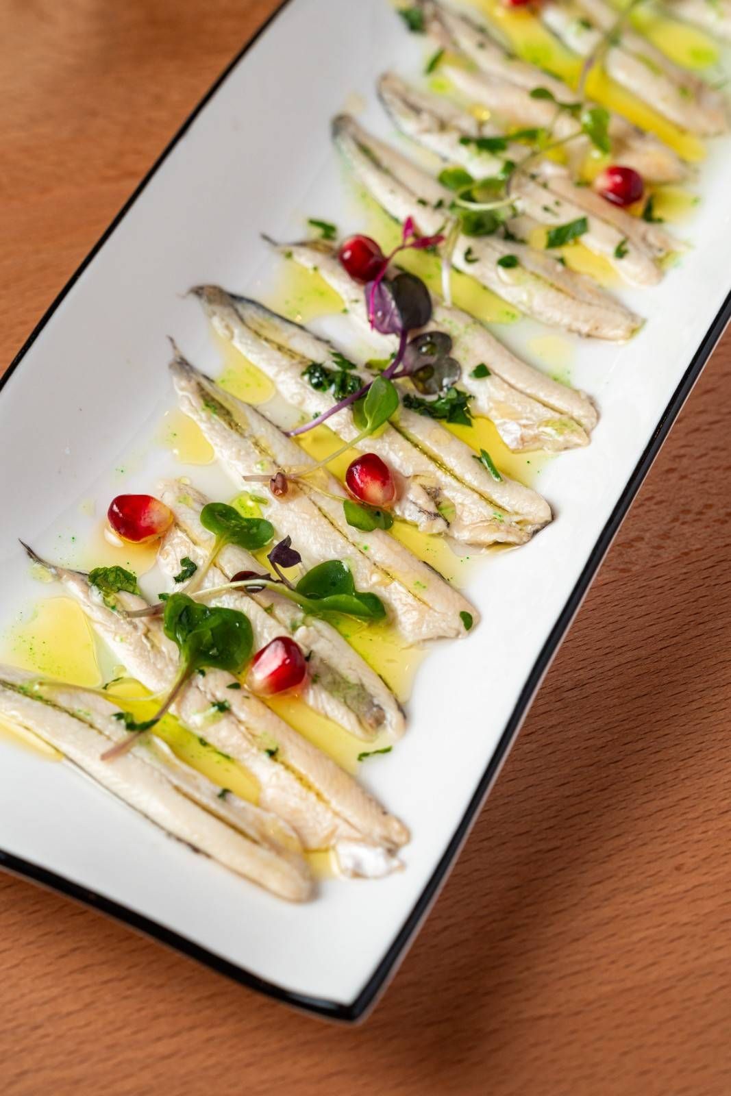 Filetes de pescado blanco emplatados, rociados con salsa, hierbas y semillas de granada en un plato rectangular.